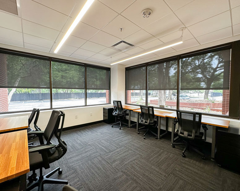 Spacious shared office with rows of desks, black chairs, and large windows.