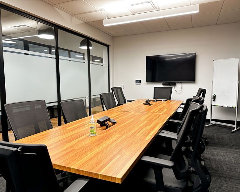 Modern conference room with a long wood table, black mesh chairs, wall-mounted TV, and whiteboard in a glass-walled space.