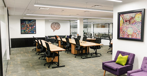 Open office with rows of wood-accented desks, black chairs, colorful wall art, and carpeted floors.